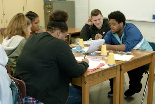 Five diverse high school students sit at a table, looking at text together and discussing as they engage in student-led team learning, a model of instruction designed to increase student achievement, close achievement gaps, and improve academic performance.