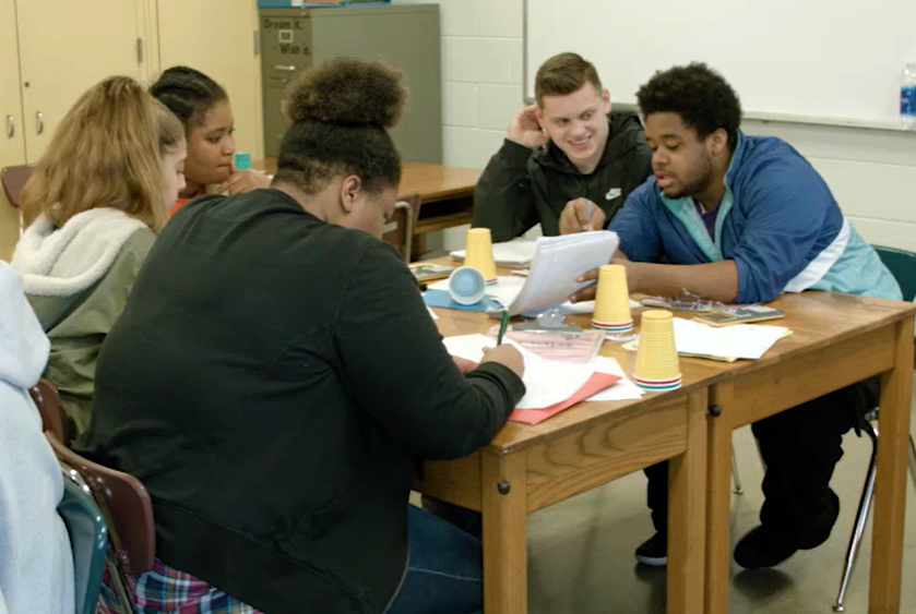 Five diverse high school students sit at a table, looking at text together and discussing as they engage in student-led team learning, a model of instruction designed to increase student achievement, close achievement gaps, and improve academic performance.