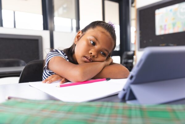 An elementary school student sits at a table with her head down staring at a screen, seemingly disengaged in the academic intervention that she has been placed in.