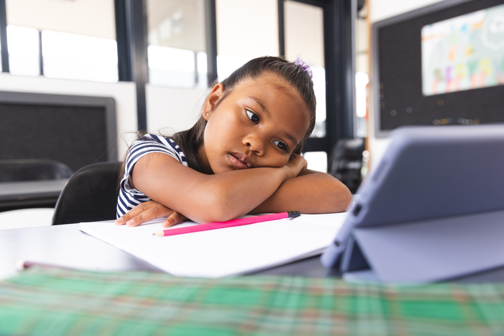 An elementary school student sits at a table with her head down staring at a screen, seemingly disengaged in the academic intervention that she has been placed in.