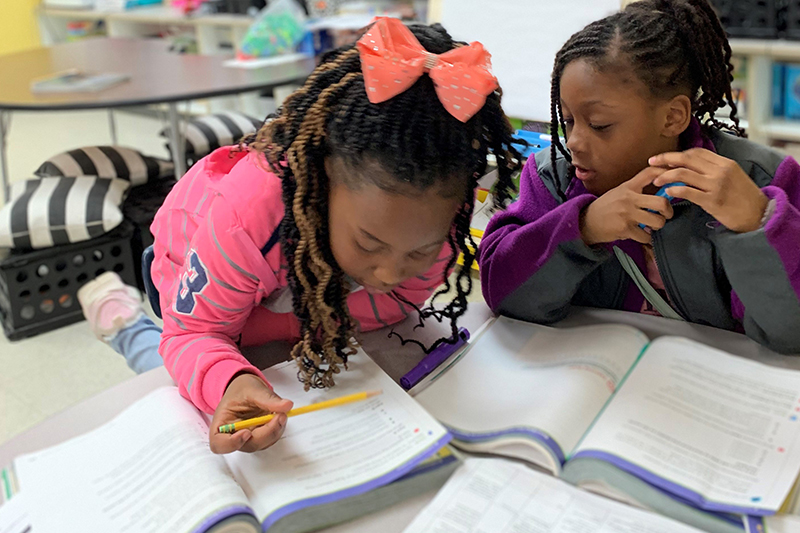 Two elementary school age students lean over a desk as they read English Language Arts curriculum resources together while leading their own learning in teams as part of the Model of Instruction for Deeper Learning.