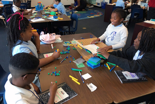 Four elementary school age students sit at a table and work as a team, holding notebooks and math manipulatives, as they use curriculum resources and lead their own learning as part of the Model of Instruction for Deeper Learning.