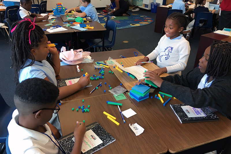 Four elementary school age students sit at a table and work as a team, holding notebooks and math manipulatives, as they use curriculum resources and lead their own learning as part of the Model of Instruction for Deeper Learning.