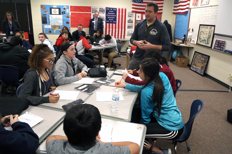 A small team of middle school aged students sit together and engage in a team task and discussion as their teacher observes as part of the Model of Instruction for Deeper Learning; students are clearly engaged and exhibiting positive behaviors.