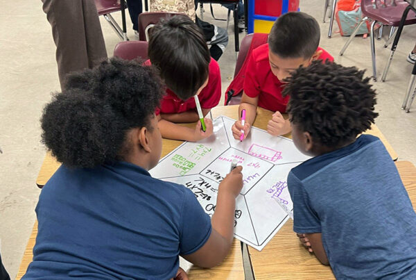 A diverse team of four elementary-aged students sit around a desk, fully engaged with positive behaviors, writing on a shared team mat resource from the Model of Instruction for Deeper Learning.