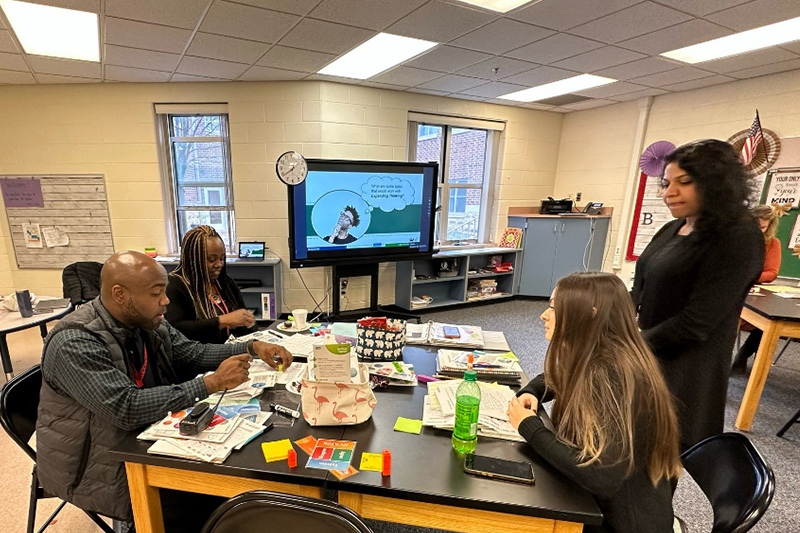 School leaders and teachers sit at a table while an expert coach looks on, guiding them through a collaborative professional development session to support school improvement after a comprehensive needs assessment. 