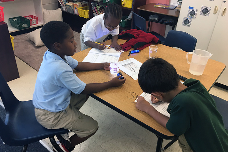 Three elementary-aged students sit at a desk doing an experiment with cups of water and small flashlights, working together as they engage in student-led team learning, a model of instruction that activates the rigor of the Next Generation Science Standards (NGSS).