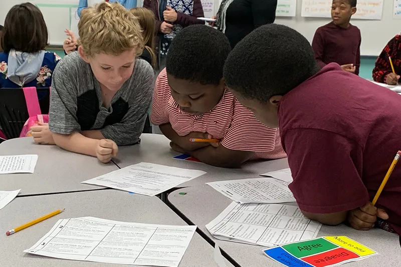 Three elementary aged students lean toward one another, fully engaged as they collaborate on their academic work, which is a powerful way to prevent chronic absenteeism. 