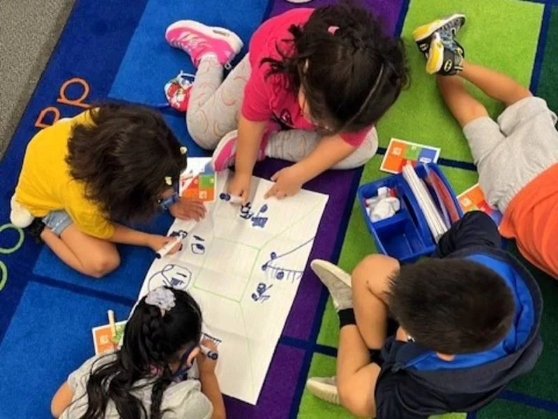 Four pre-K students sit together on a carpet, writing on a Summarizing Mat from the Model of Instruction for Deeper Learning as they develop early literacy skills and experience the full rigor of the pre-K curriculum.