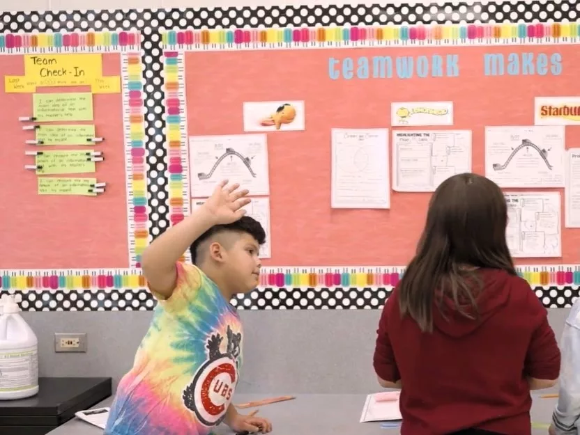 Two elementary students stand in front of a board that has student work and the words “teamwork makes the dream work” as they engage in rigorous academic discourse.