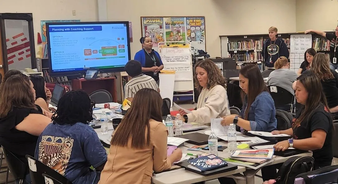 About a dozen teachers and leaders sit in a classroom together in groups as they engage in professional development on a research-based instructional model that reduces teacher burnout.