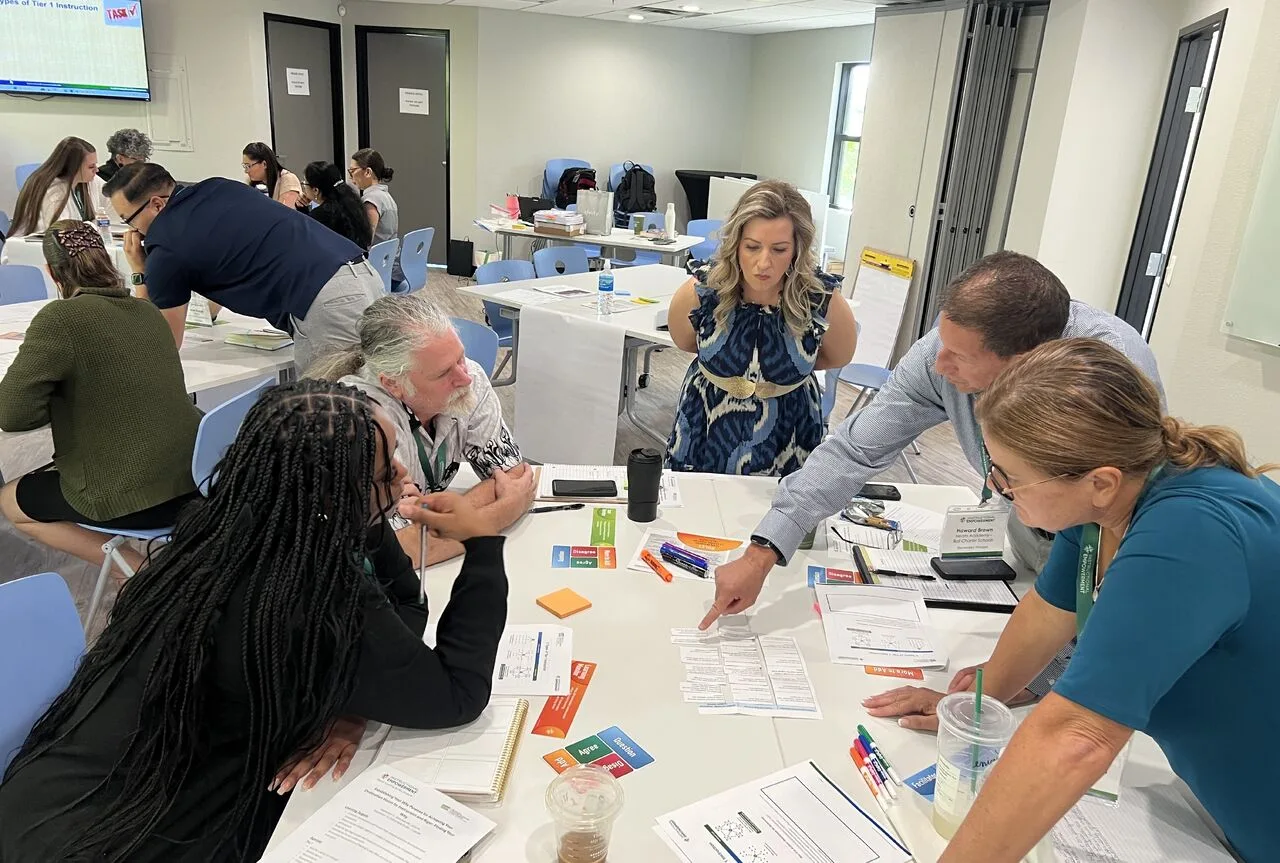 Five education leaders (superintendents, district leaders, principals) stand and sit around a table where they are engaged in professional development and coaching on how to improve Tier 1 instruction, one of the market share systems that increases student enrollment.