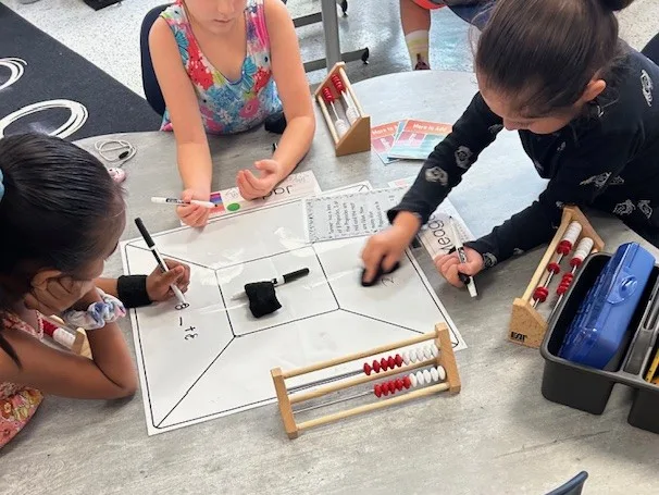 Three students write on a Summarizing Mat from the Model of Instruction for Deeper Learning and use math manipulatives.