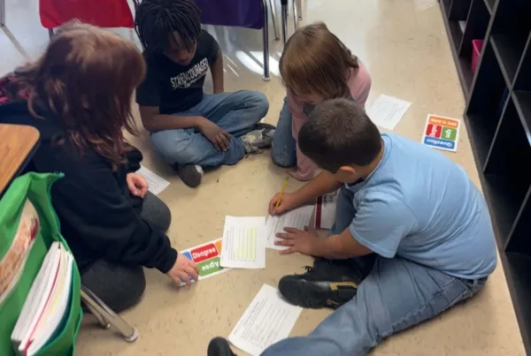 Four elementary age students sit on the floor of the classroom, working collaboratively as they write and use Agree/Disagree cards from the Model of Instruction for Deeper Learning, which increases academic rigor and can be monitored via walkthroughs using the RigorWalk tool to ensure schools are on track to raise achievement.