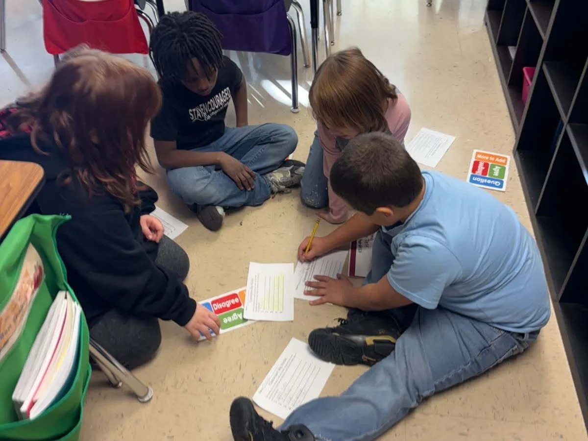 Four elementary age students sit on the floor of the classroom, working collaboratively as they write and use Agree/Disagree cards from the Model of Instruction for Deeper Learning, which increases academic rigor and can be monitored via walkthroughs using the RigorWalk tool to ensure schools are on track to raise achievement.