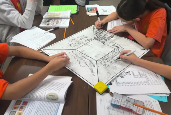 Four elementary students from a turnaround school sit at a table together working on a history lesson and writing on a mat as they collaborate in rigorous teams as part of the research-validated Model of Instruction for Deeper Learning.
