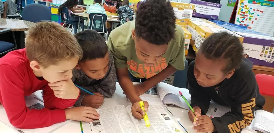 Four students from Moseley Elementary, featured in a turnaround school case study, lean over a table together with highlighters and pencils as they collaborate in a rigorous team as part of the Model of Instruction for Deeper Learning.