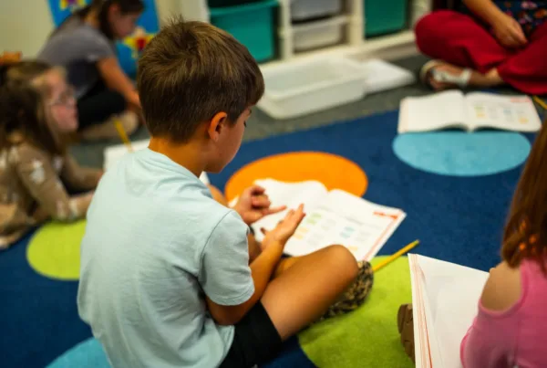 Elementary students sit on a classroom carpet in Colorado Springs School District 11, working from printed high-quality instructional materials during a student-led team lesson.